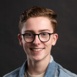 Lachende jongeman met bril op en spijkerjack aan, tegen een zwarte achtergrond